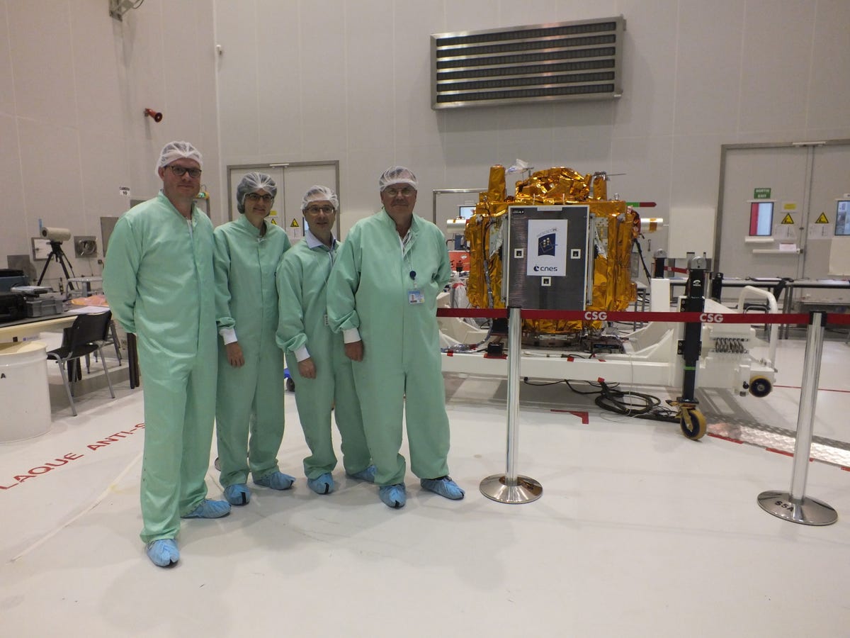 Vier Wissenschaftler, gekleidet in grüne Kleidung und Haarnetze, stehen neben einem ofengroßen Gerät, das in Goldfolie eingewickelt ist.